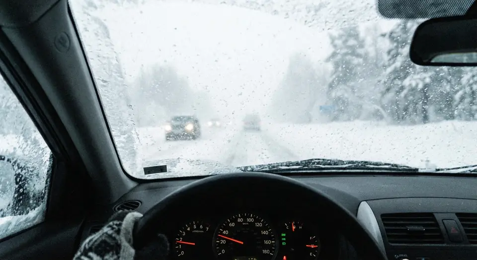 A view through a frosty windshield of a snowy road, with the dashboard in the foreground and another car ahead.